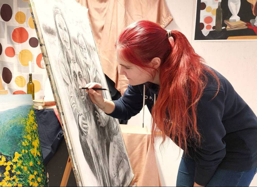 Opiskelija maalaa rantaviivassa istuen, toinen opiskelija kahlaa taustalla